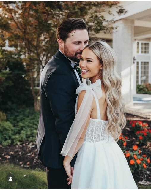 Couple in wedding attire smiling outdoors, surrounded by greenery.
